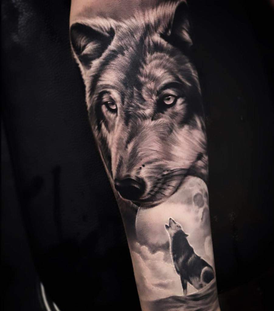 Tattoo of a wolf's face and a howling wolf beneath a full moon in grayscale on an arm