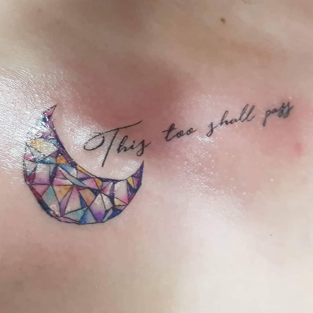 A "This Too Shall Pass" tattoo on a person's upper chest, accompanied by a colorful, geometric crescent moon design.