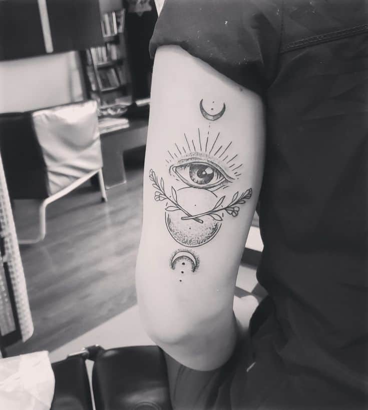 Tattoo of an all-seeing eye with crescent moons and leaves, inked in fine black lines on the upper arm.