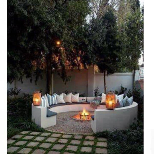 Circular outdoor seating area with fire pit, pillows, and lanterns surrounded by trees.