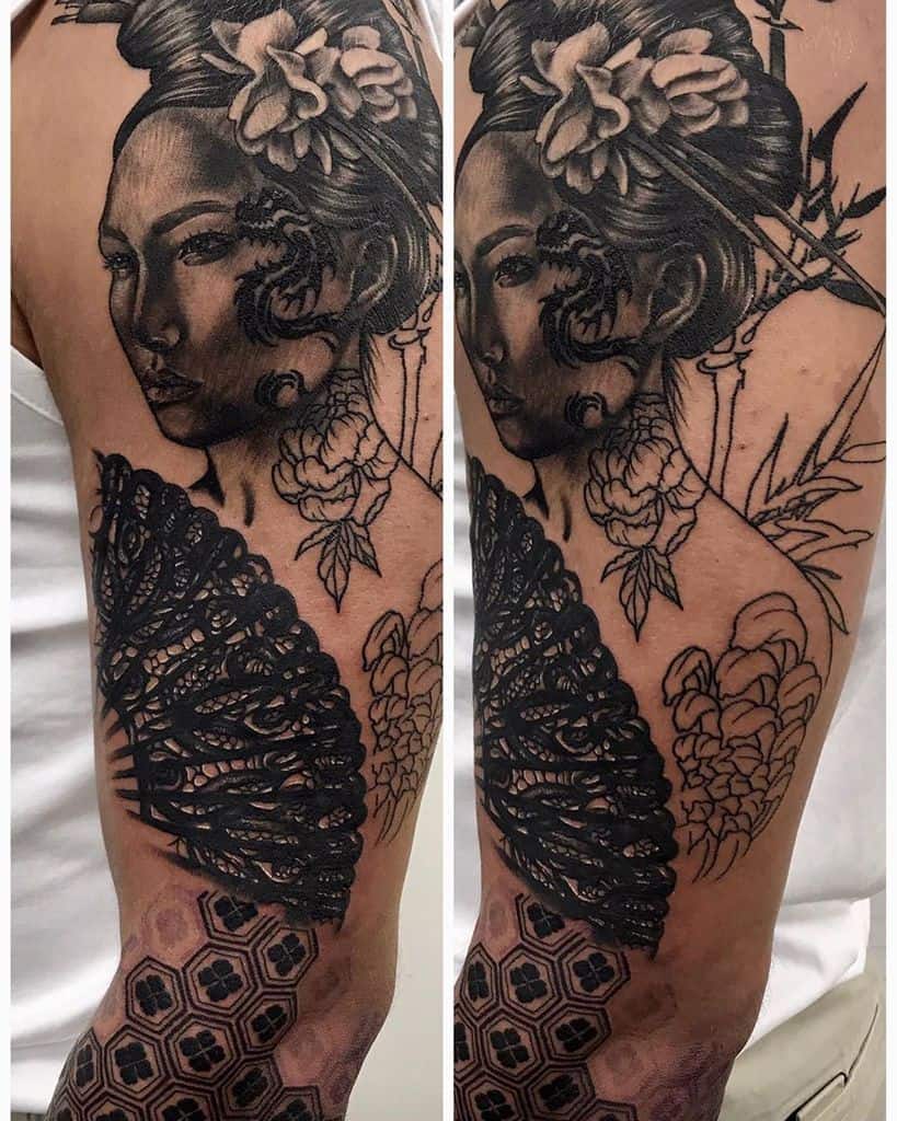 Black and gray upper arm tattoo of a Geisha with floral hair ornaments, intricate shading, a bold lace-patterned fan, and geometric forearm designs.