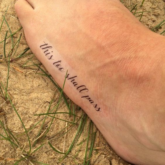 A "This Too Shall Pass" tattoo in cursive on the top of a person's foot, with sand and grass around it.
