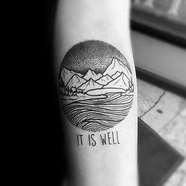 Black ink circular tattoo featuring mountains, waves, and dot shading, with the phrase "It Is Well" beneath, symbolizing peace and resilience.