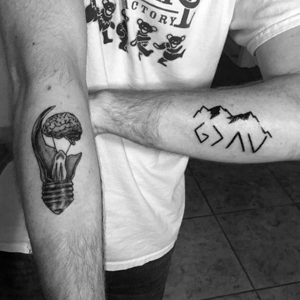 Dual forearm tattoos featuring "God is greater than the highs and lows" symbol with mountains on one arm and a creative brain-lightbulb design on the other arm