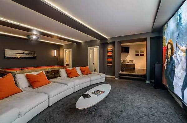Modern media room with a large screen, white sectional sofa, orange pillows, and a pool table in the background