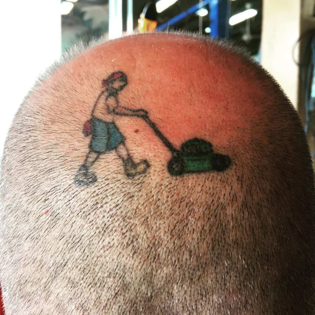A humorous tattoo on a bald head featuring a tiny person pushing a lawnmower, creating the illusion of mowing the scalp.