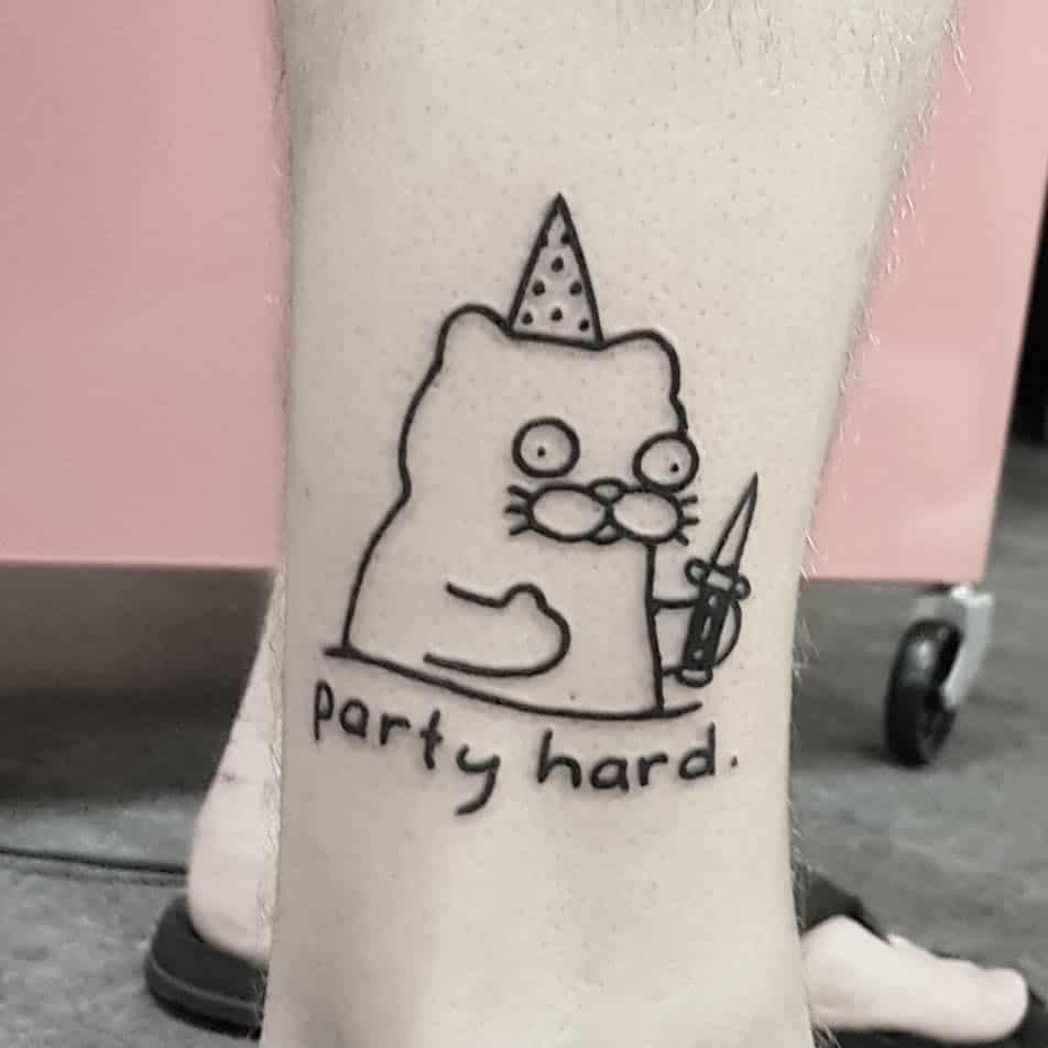 A black ink tattoo of a wide-eyed cat wearing a party hat, holding a knife, with the phrase 'party hard.' in a humorous and ironic style.