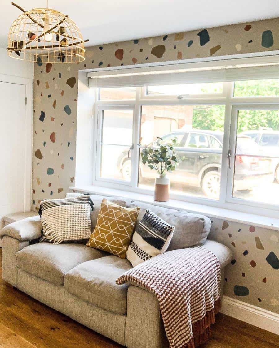 Cozy living room with a beige couch, accent wall, patterned pillows, and terrazzo wallpaper. Large window and rattan light fixture