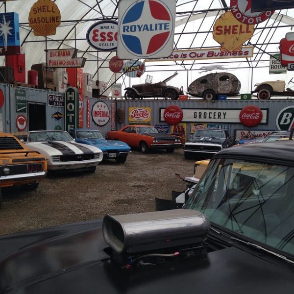 Rustic garage showcasing muscle cars and vintage automotive signs under a greenhouse-style roof.