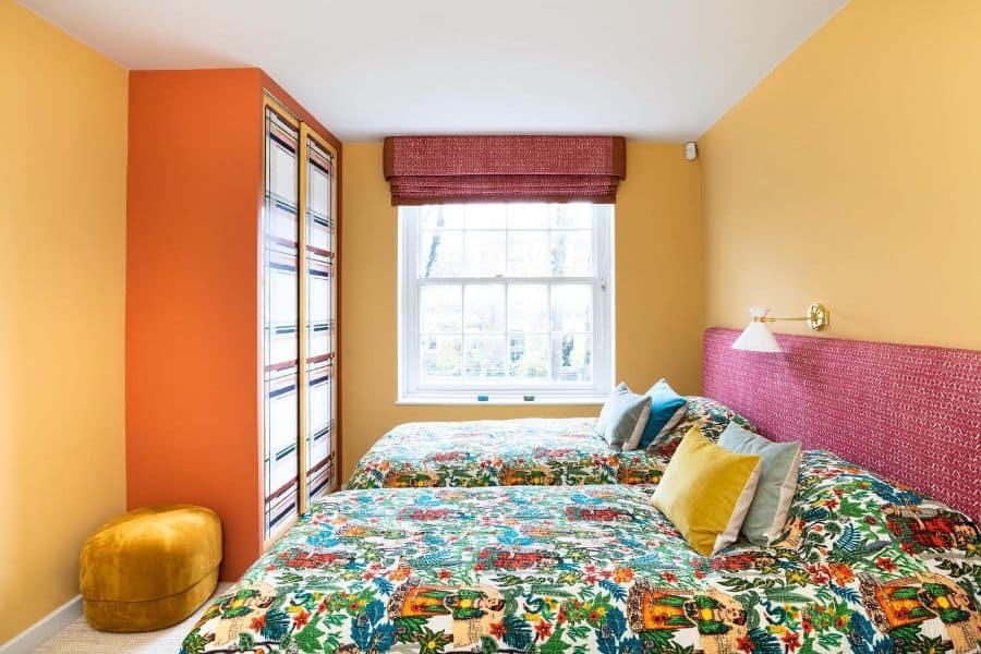 Colorful bedroom with two patterned beds, orange walls, yellow window valance, and a mustard pouf