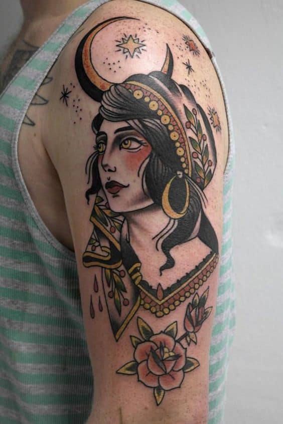 Traditional tattoo of a gypsy woman with a crescent moon, gold jewelry, and floral details, featuring bold shading on the upper arm.