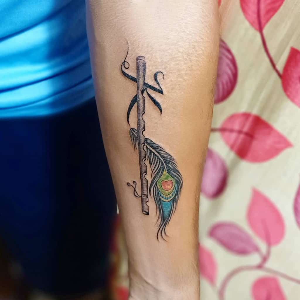 Forearm tattoo featuring a wooden flute with a tied peacock feather, detailed shading, and vibrant blue, green, and gold hues in the feather.
