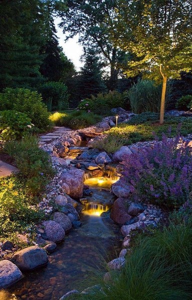 Beautiful sloped garden with a natural rock waterfall, soft lighting, lush greenery, and blooming plants creating a serene evening landscape