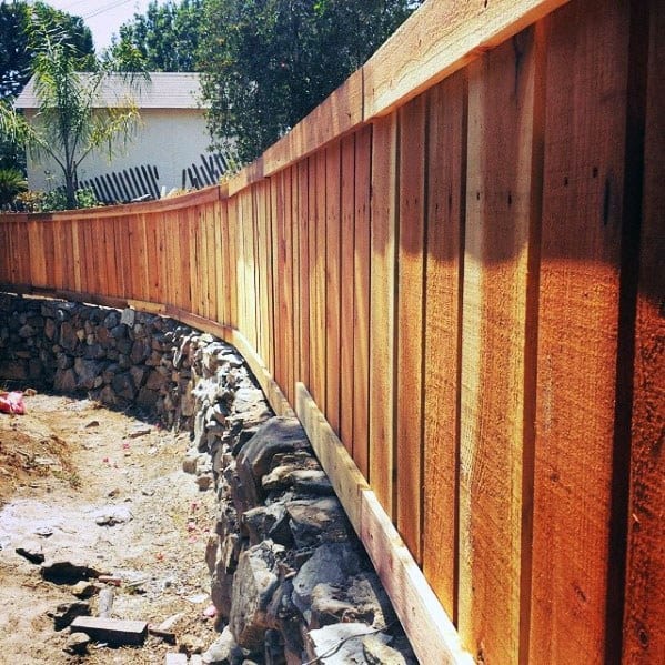 Curved wooden fence crowns a stone wall, with a dirt path and trees in the background
