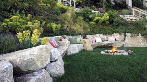 Natural stone retaining wall with built-in seating, decorative cushions, and a fire pit on a lawn.