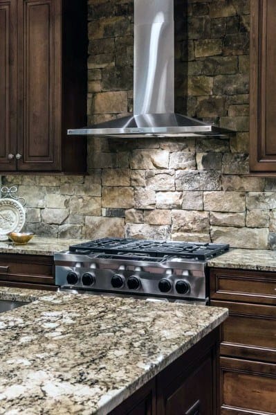Rustic kitchen with a natural stone backsplash, rich wood cabinetry, stainless steel range hood, and granite countertops