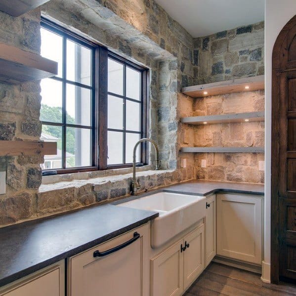 Rustic kitchen with natural stone walls, open wooden shelves, farmhouse sink, and large windows for natural light