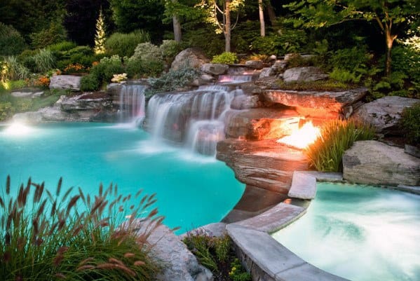 Illuminated backyard pool with cascading waterfall, surrounded by rocks and lush greenery