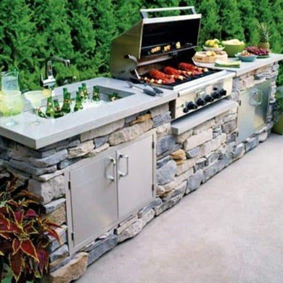 Outdoor kitchen with built-in grill, sink, fruit, salads, and drinks surrounded by lush greenery