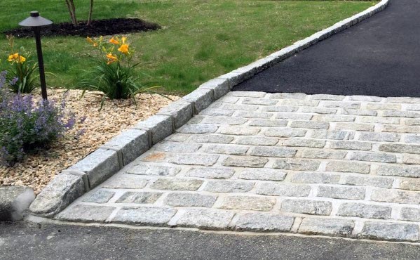 Asphalt driveway with natural stone edging and a cobblestone threshold next to a landscaped garden.
