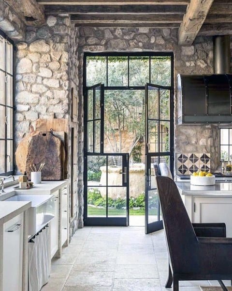 Rustic kitchen with stone walls, metal-framed glass doors, white cabinets, and garden views.