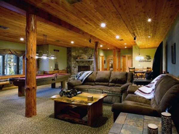 Rustic basement with natural wood beam poles, pool table, and stone fireplace.