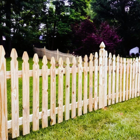 A charming wooden picket fence graces the grassy yard, with trees and a house creating a picturesque backdrop