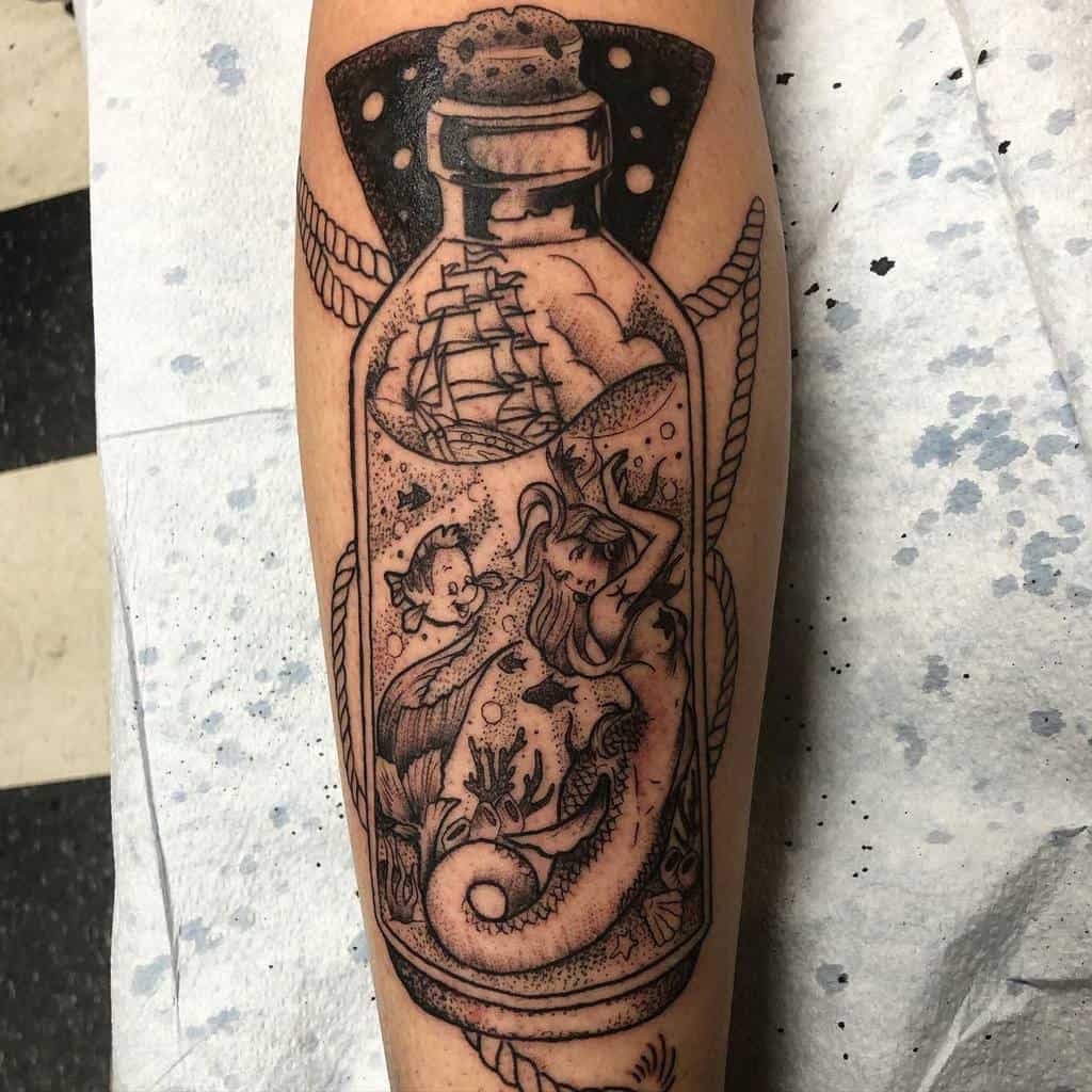 Black and grey tattoo of a mermaid inside a bottle, surrounded by underwater elements, with a sailing ship at the top of the design.