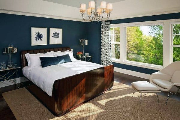 Spacious navy bedroom with wooden bed frame, chandelier, and large window with natural light.