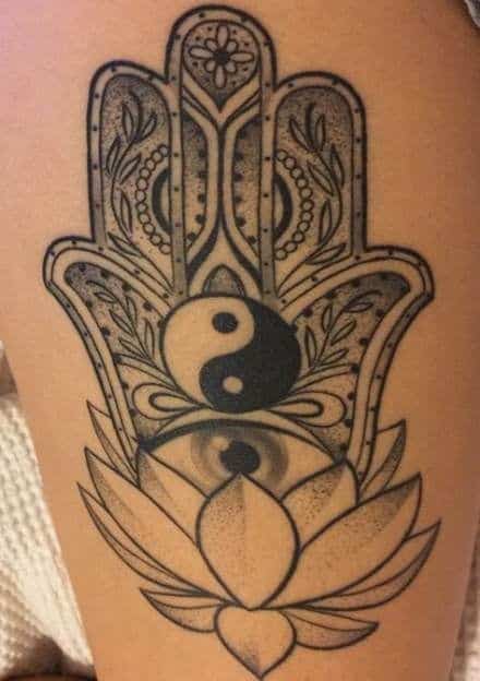 Tattoo of a Hamsa hand with intricate patterns, a yin-yang symbol, an all-seeing eye, and a blooming lotus, inked in black with dotwork shading.