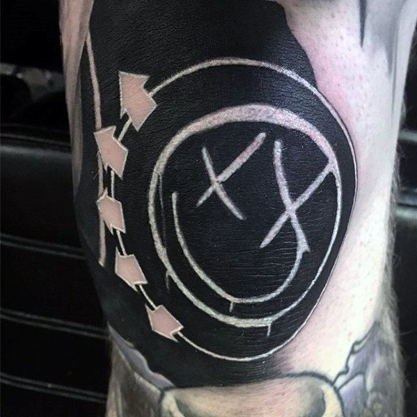 Black and gray tattoo of the Blink-182 smiley face logo with bold white arrows, placed on the forearm, giving it a striking contrast with detailed shading.