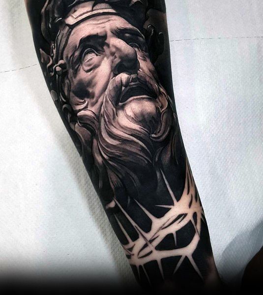 Realistic black and grey tattoo of a classical bearded statue with dramatic lighting, symbolizing wisdom, strength, and timeless divinity.
