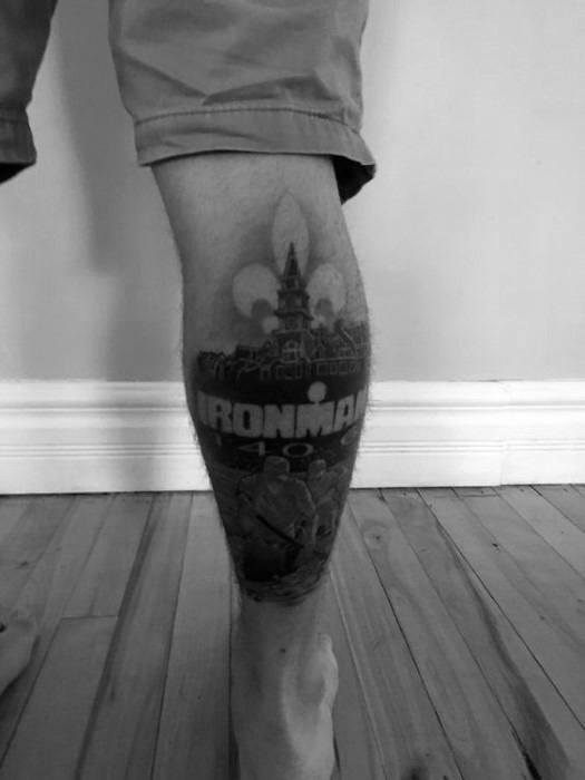 Black and grey tattoo featuring the Ironman logo with a detailed background of a cityscape and a fleur-de-lis, symbolizing achievement and personal connection to the race.