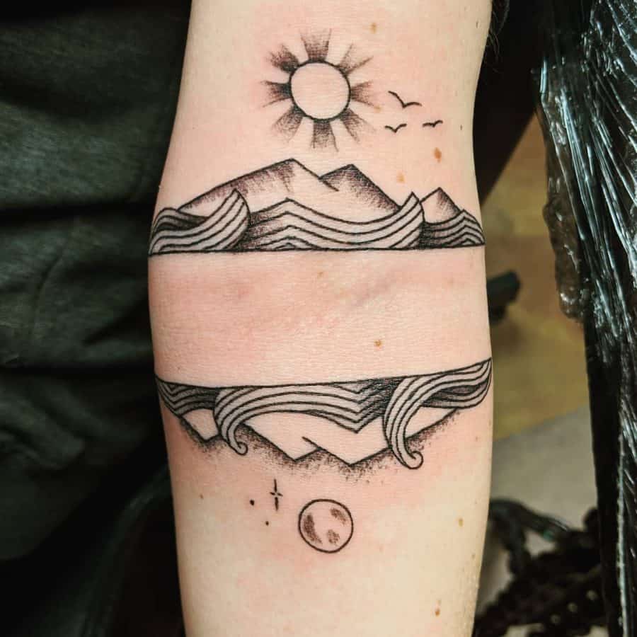 Black ink tattoo of a landscape split by a negative space band, featuring mountains, waves, a sun with birds above, and a moon with stars below.
