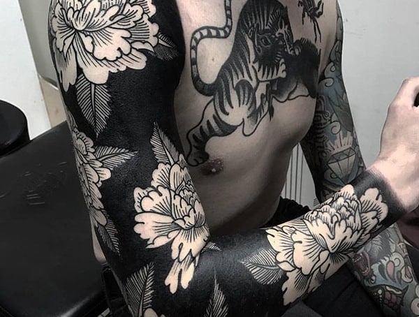 Blackout tattoo sleeve featuring negative space floral designs, creating a bold contrast with intricate flowers against a dark background.