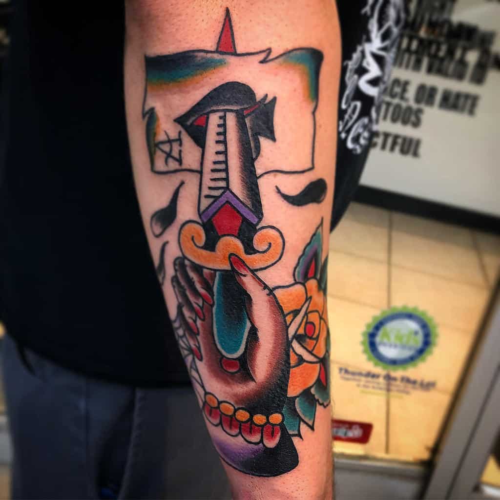 A colorful tattoo of a hand holding a dagger, stabbing the Ace of Spades card, surrounded by droplets and a rose design