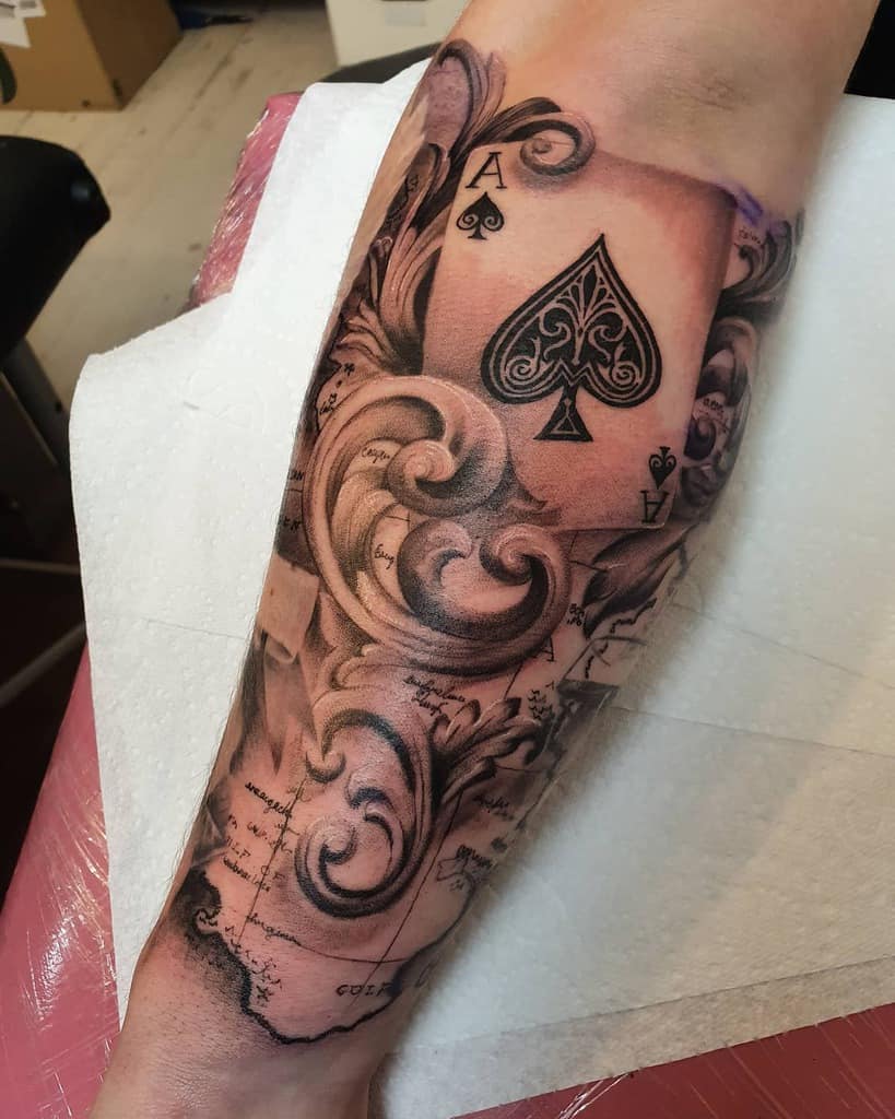 A black-and-white tattoo of the Ace of Spades card, surrounded by ornate swirls and intricate shading and a map background
