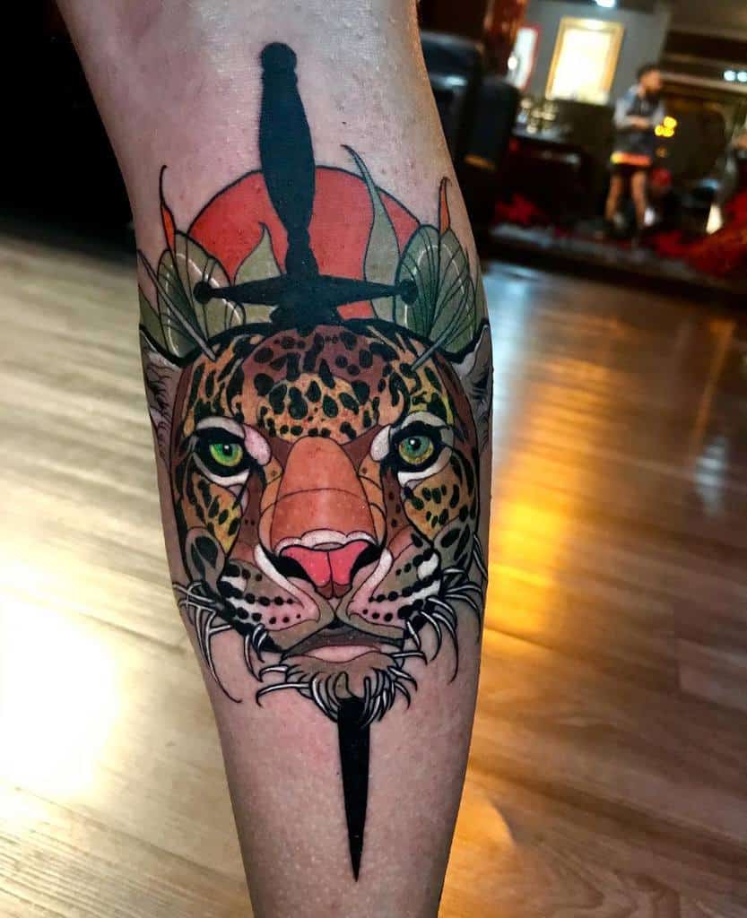 A vibrant tattoo of a jaguar's face, surrounded by colorful accents and a bold knife through the center, placed on the forearm with striking details and dynamic colors.