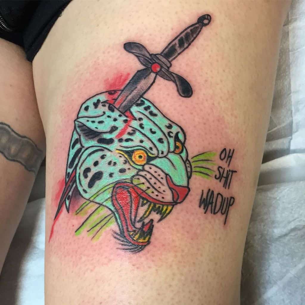 A bold and colorful tattoo of a snarling jaguar with a sword through its head, featuring bright turquoise and red tones, with playful text reading "OH SHIT WADUP" on the thigh.
