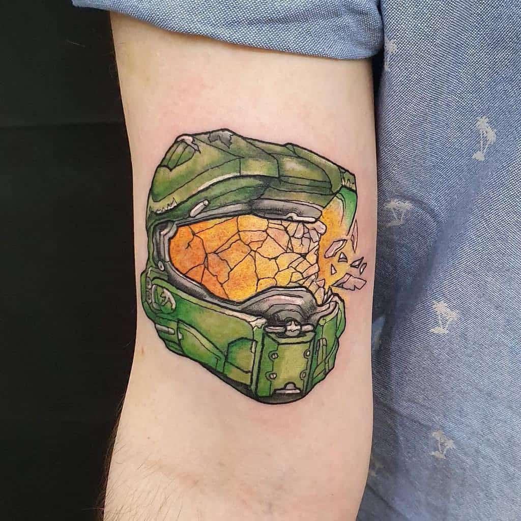 Detailed Halo tattoo of Master Chief’s cracked helmet, featuring vibrant green armor and a shattered golden visor, inked on the upper arm.