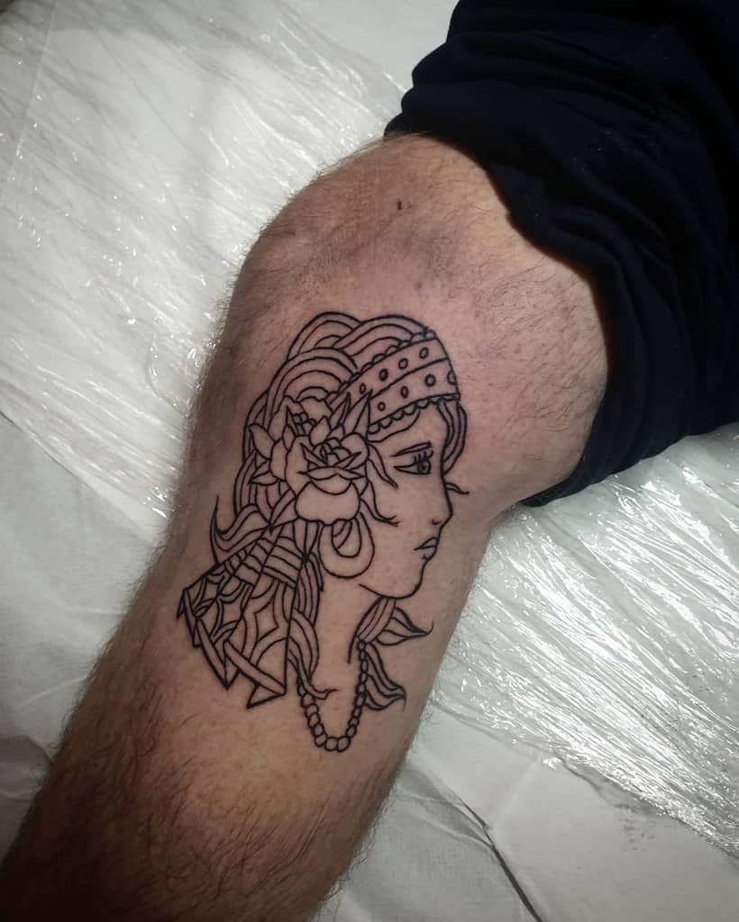 Linework tattoo of a gypsy woman in profile with a headscarf, rose, and jewelry on the leg, featuring bold black outlines.