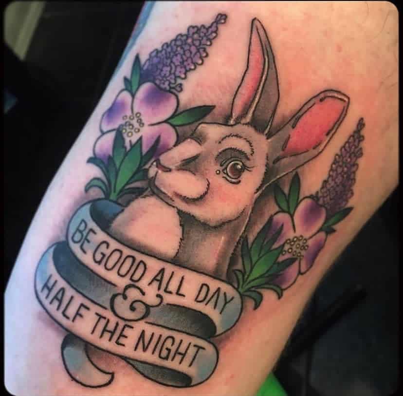 Traditional-style tattoo of a rabbit surrounded by lilac flowers and a banner reading 'Be good all day & half the night'.