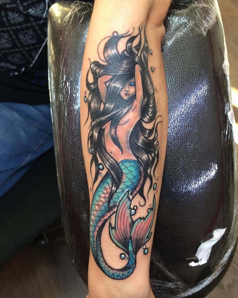 Colorful tattoo of a mermaid with long flowing black hair, blue eyes, and a vibrant turquoise and pink tail, surrounded by water bubbles.