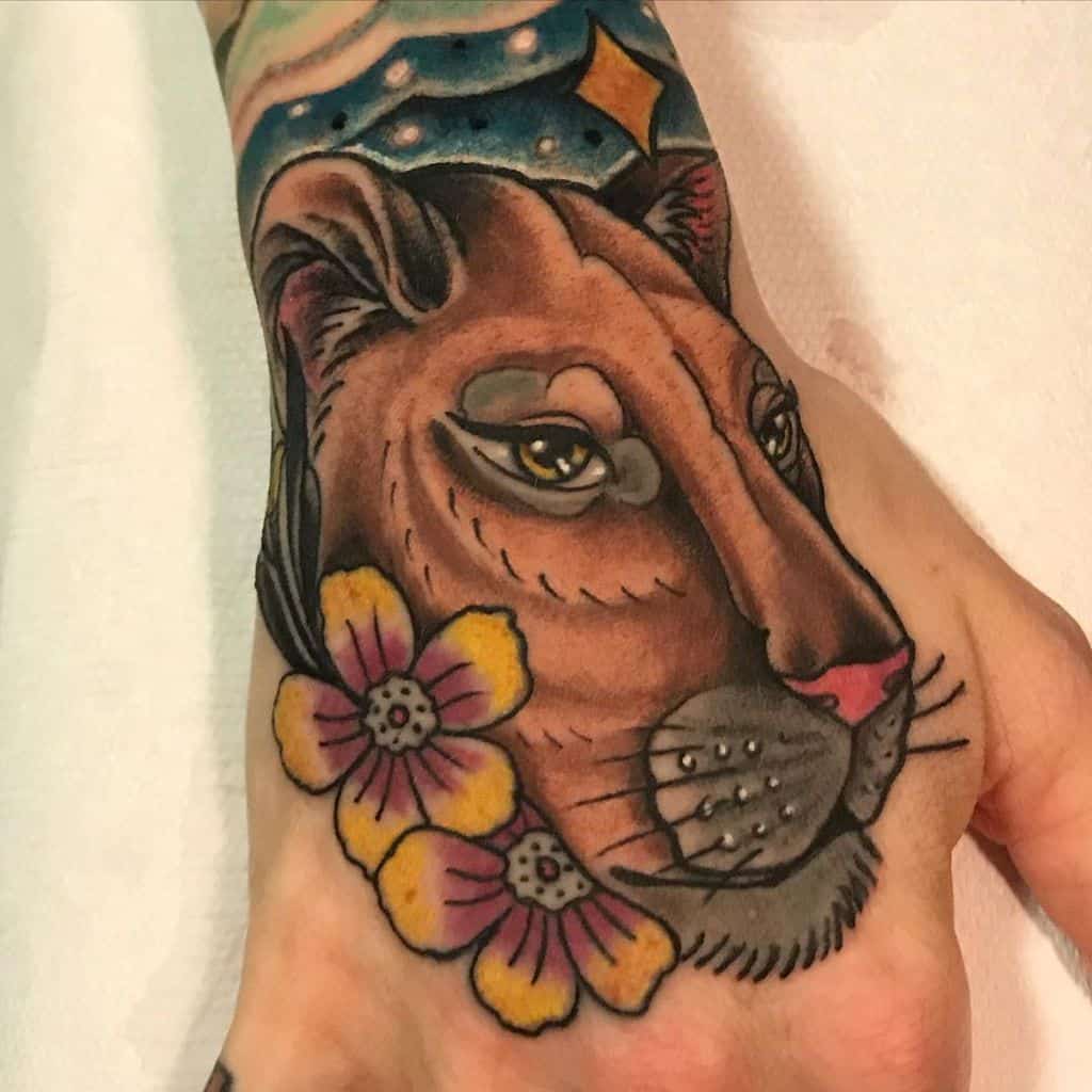 Hand tattoo of a lioness with expressive eyes, yellow flowers, and a decorative design on the wrist in bold, colorful ink.