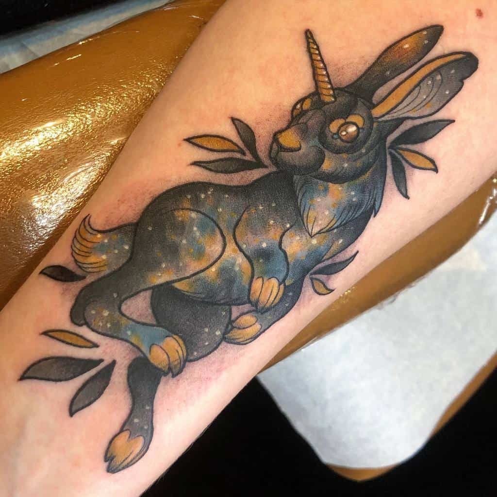 Whimsical tattoo of a rabbit with a unicorn horn, surrounded by soft glowing galaxy-like colors and leaves, with vibrant blues and golds