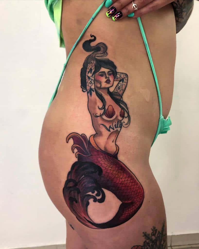 Neo-traditional hip tattoo of a siren with vibrant red tail, shell accents, and 'Wild' script, blending bold and delicate details