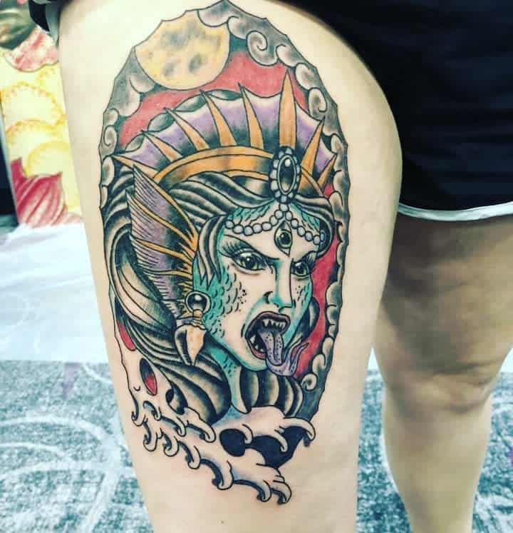 Colorful tattoo of a mythical siren with a crown, fangs, and waves on a thigh