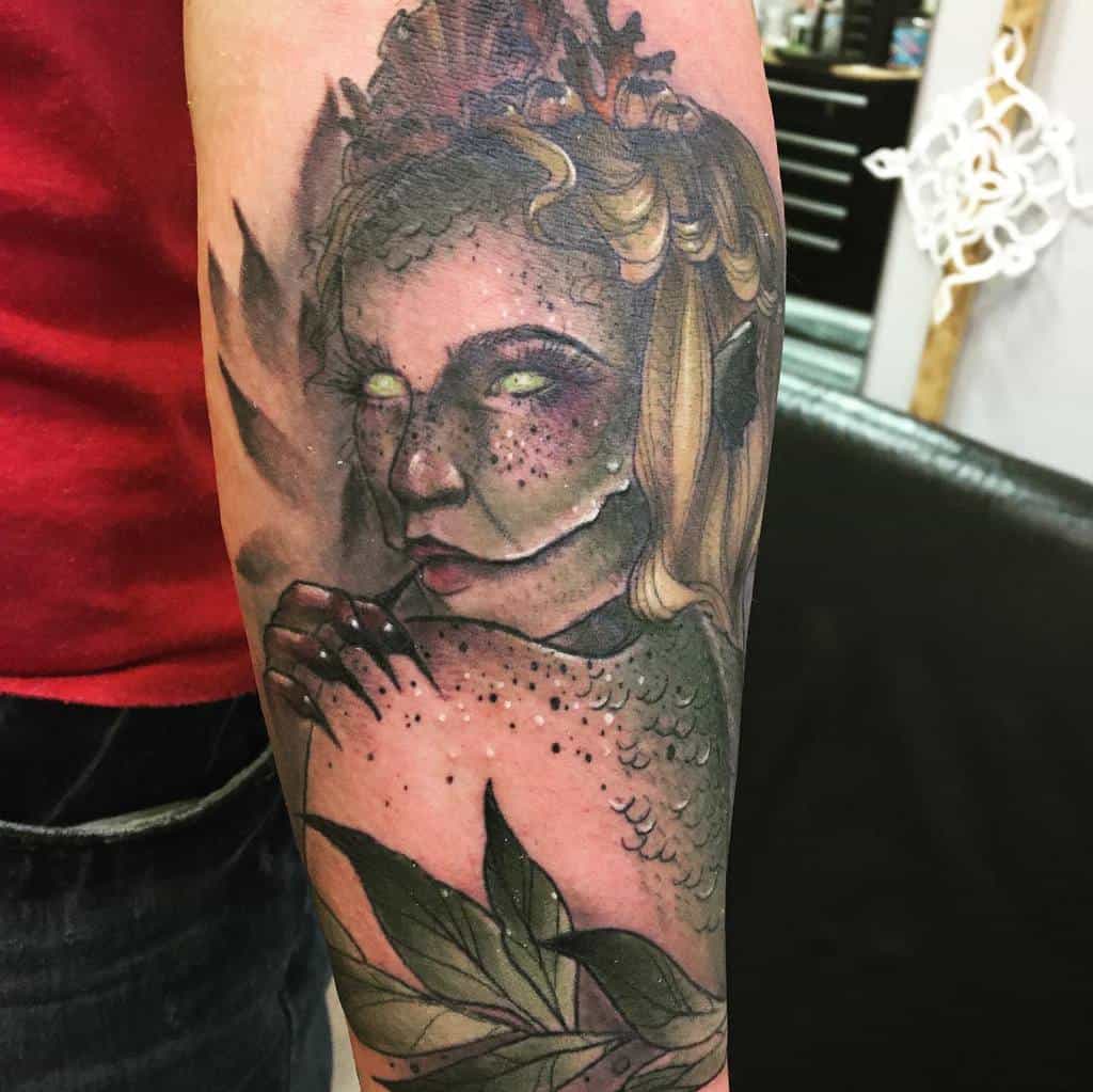 Moody neo-traditional tattoo of a siren with glowing eyes, green scales, and intricate leafy details on the arm