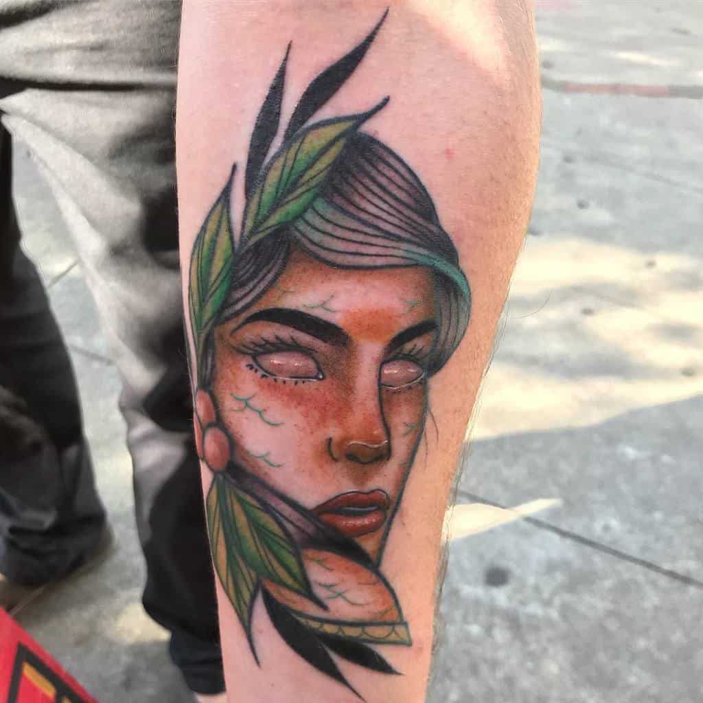 Neo-traditional tattoo featuring a mystical female figure with glowing white eyes, surrounded by green leaves, adding an ethereal and nature-inspired touch