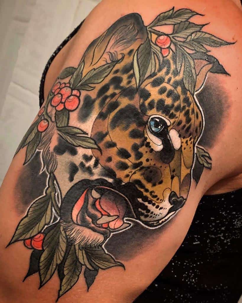 A detailed tattoo of a jaguar's face with vivid blue eyes, surrounded by lush green leaves and red berries, placed on the upper arm for a bold and natural design.
